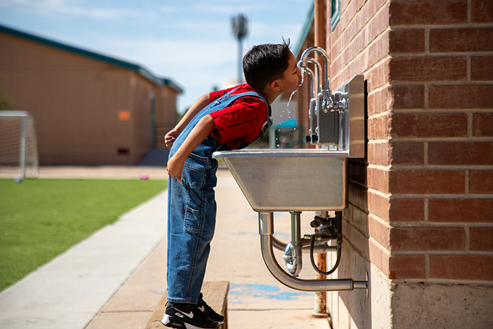 A boy stands on a stool to drink water from an outdoor sink