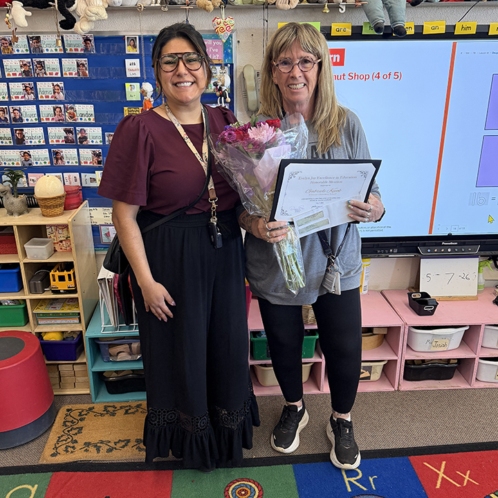 A woman, right, holds up an award certificate and flowers next to another woman